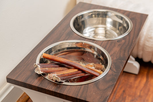 Two stainless steel dog bowls set in a dark wooden stand, one filled with bully sticks and the other empty.