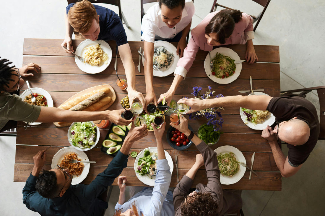 A group of people sitting around a wooden dining table sharing a meal and raising glasses in a toast, with plates of food, salads, and bread spread out in front of them.