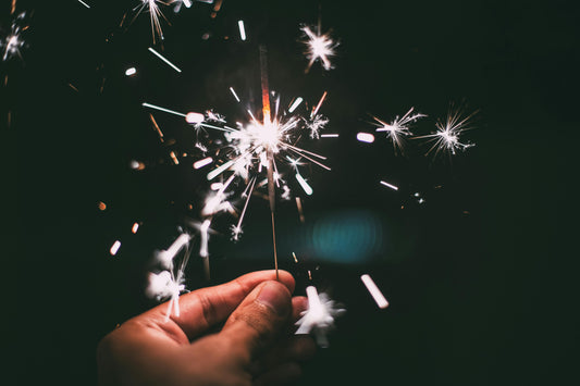 A hand holding a lit sparkler in the dark, with bright sparks glowing and spreading light against a black background.