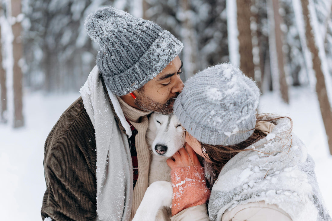 A couple wearing winter hats and scarves lovingly hugging and kissing their white dog in a snowy forest.