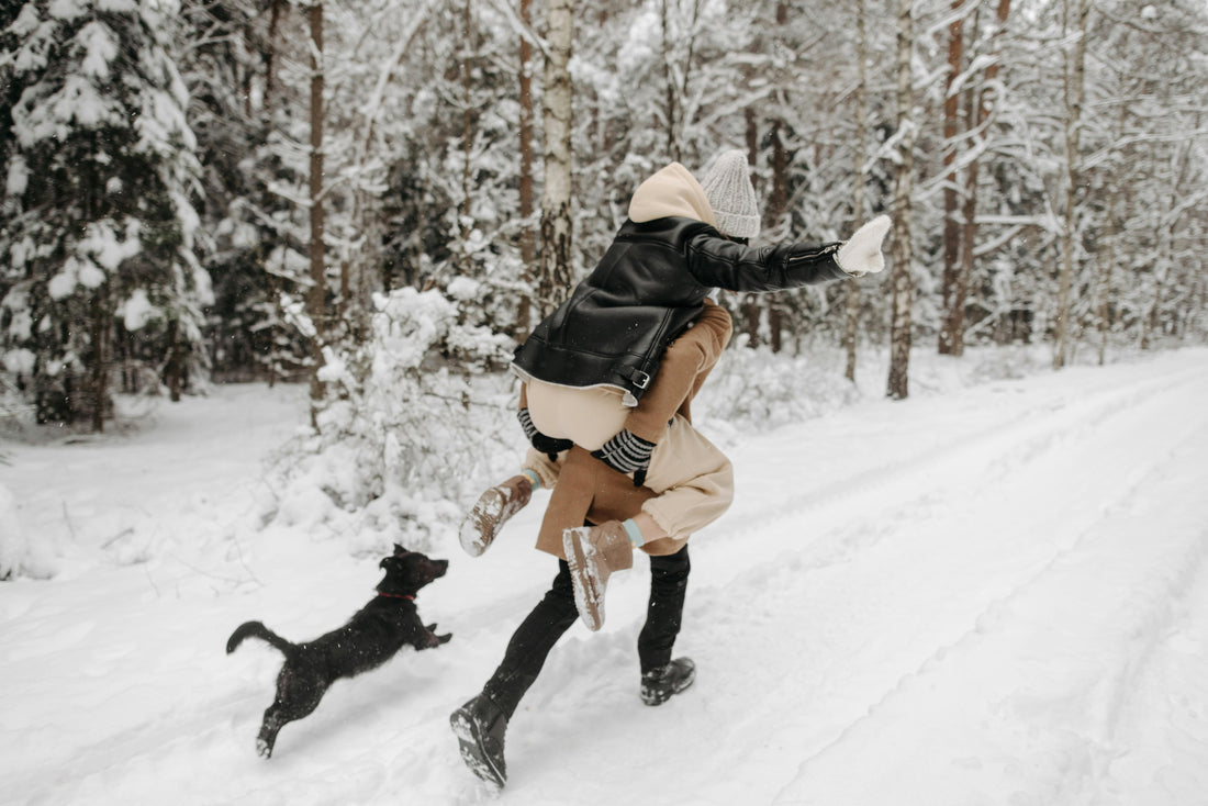 A person playfully carrying another on their back while running through a snowy forest, with a small black dog joyfully chasing beside them.