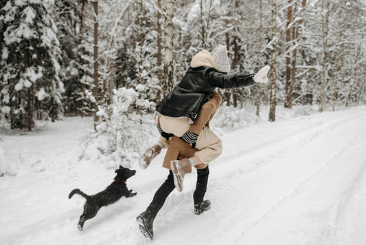 A person playfully carrying another on their back while running through a snowy forest, with a small black dog joyfully chasing beside them.