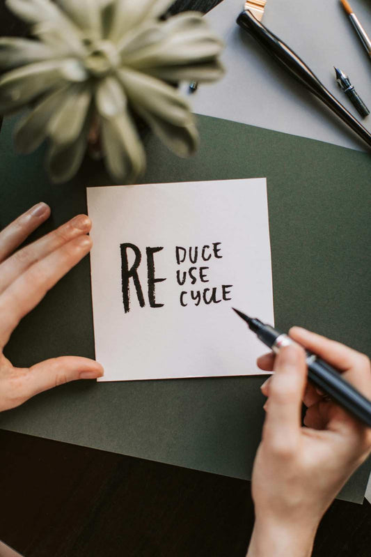 Person writing the words “Reduce Reuse Recycle” on a white piece of paper with a black marker, placed on a green surface near a plant and pens.