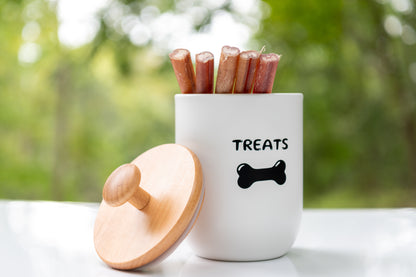 White container labeled 'TREATS' with wooden lid, filled with bully sticks, on a blurred natural background.