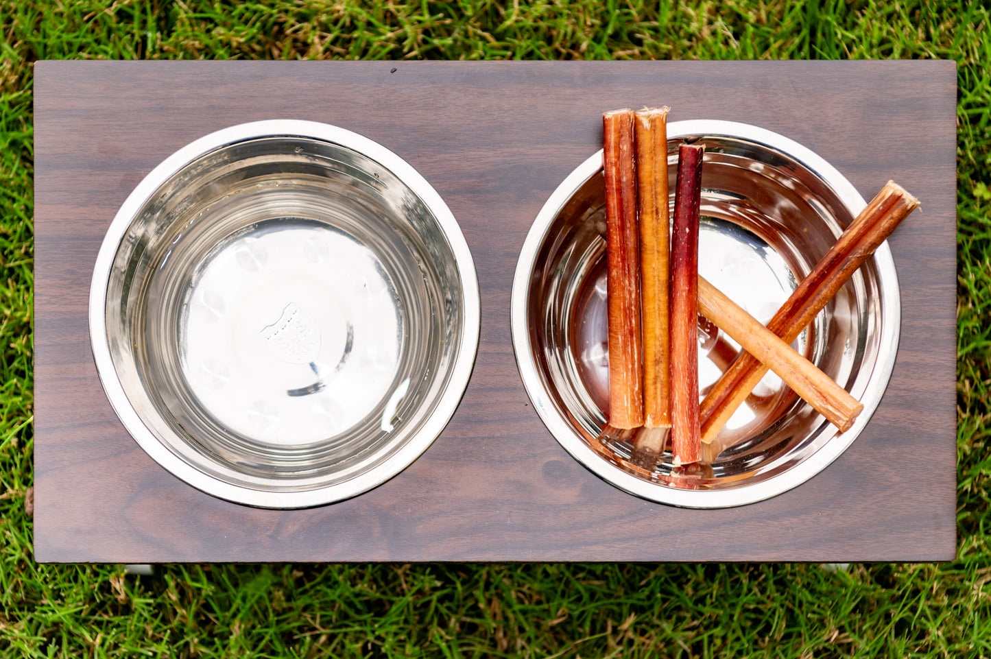 Two metal bowls on a wooden platform with one containing five straight bully sticks, set against a grass background.